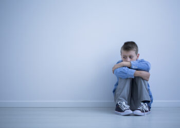 Alienated boy sitting alone on floor against wall with copy space. Sad child with autism concept